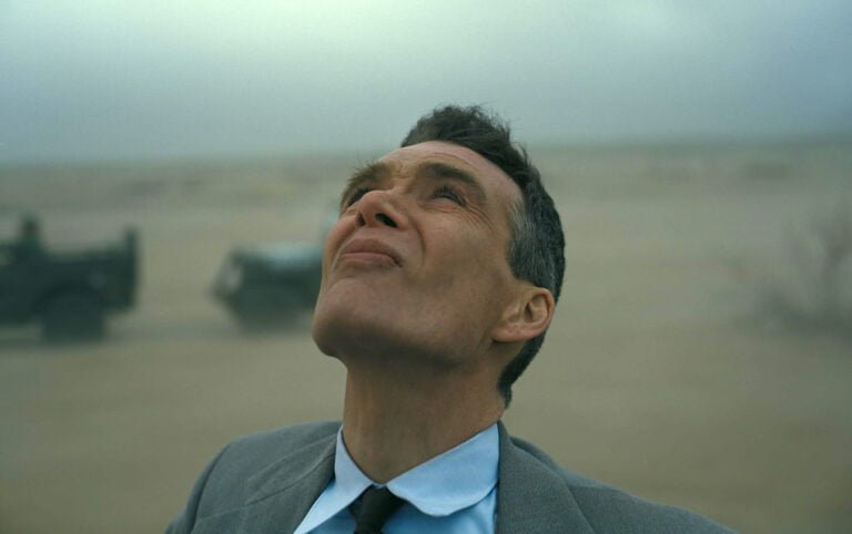 Mężczyzna w garniturze i krawacie patrzy w górę z zamyśloną miną na tle mglistej pustyni z niewyraźnym pojazdem wojskowym w tle. Kadr z filmu Oppenheimer który pojawi się na SkyShowtime.