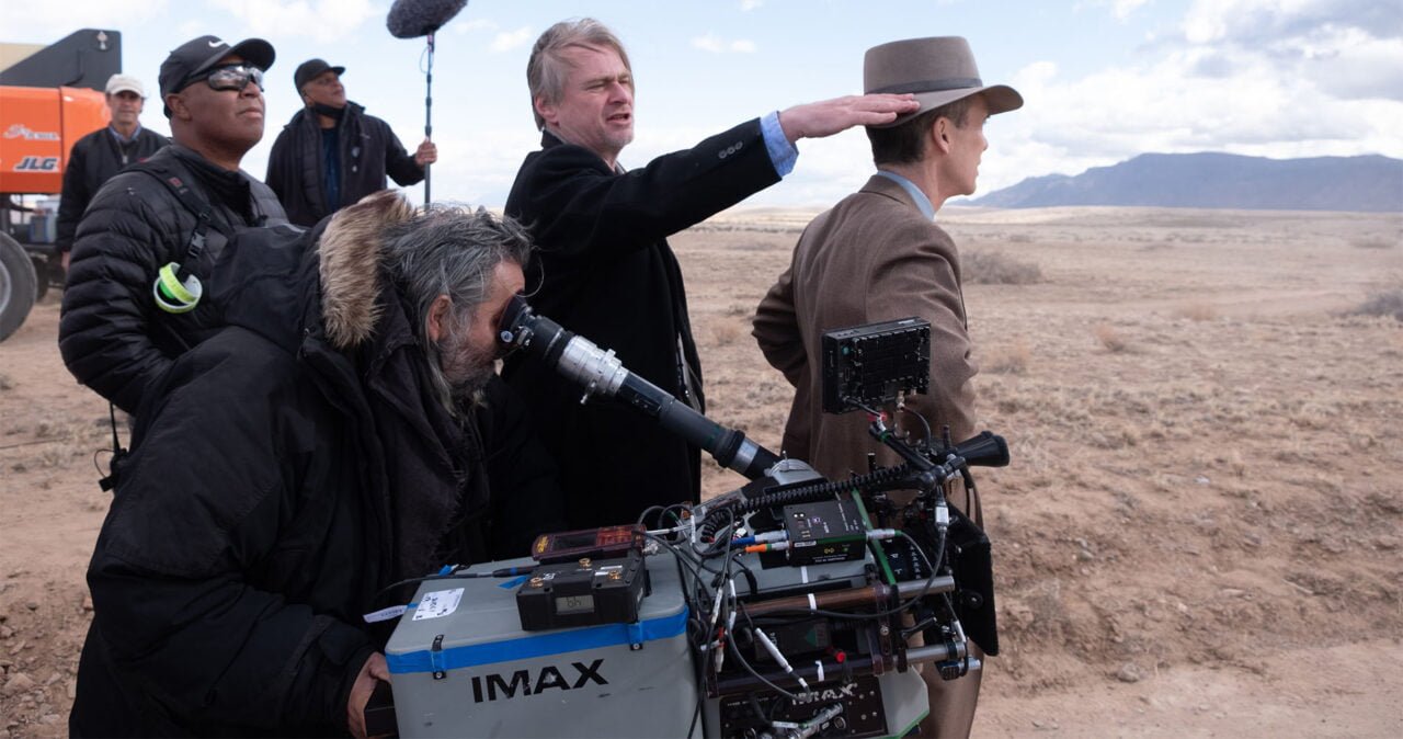 Zdjęcie przedstawia ekipę filmową na planie zewnętrznym, w tym operatora kamery IMAX, reżysera dającego wskazówki i aktora w kostiumie, w tle widoczna pustynia.