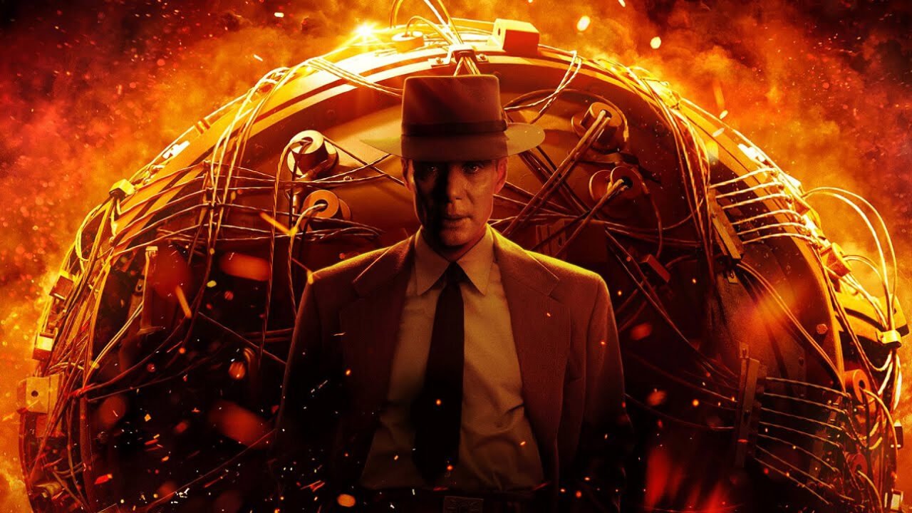 Oppenheimer film. Mężczyzna w brązowym garniturze i kapeluszu stoi na tle skomplikowanej konstrukcji przypominającej klatkę z fragmentami iskrzących się przewodów, otoczonej przez czerwonawo-pomarańczową poświatę i unoszące się iskry.