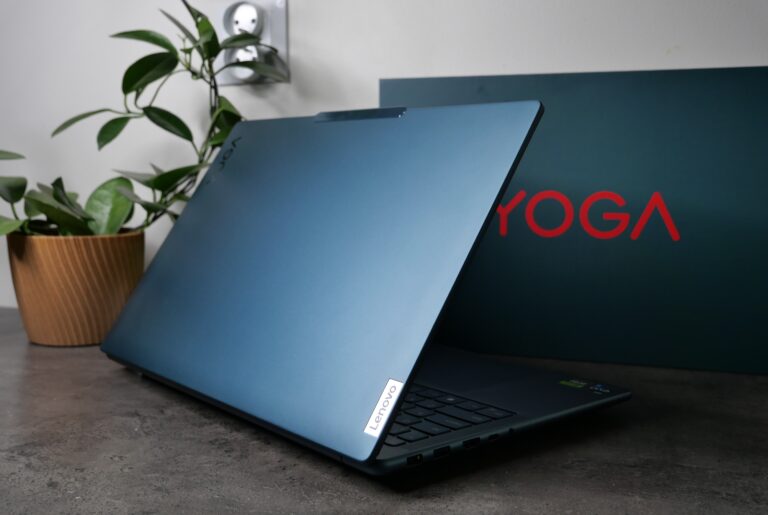Niebieski laptop Lenovo na biurku z logotypem YOGA w tle i doniczką z rośliną po lewej stronie.