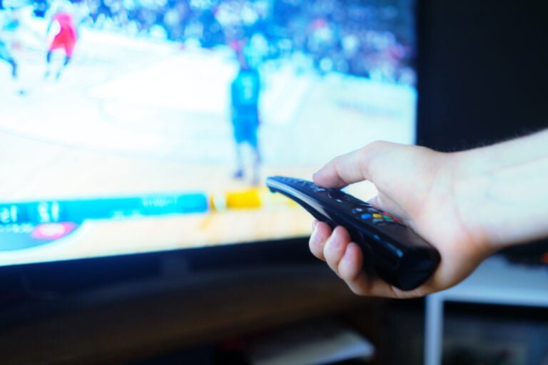 Ręka trzymająca pilota do telewizora z rozmytym obrazem meczu koszykówki w tle.