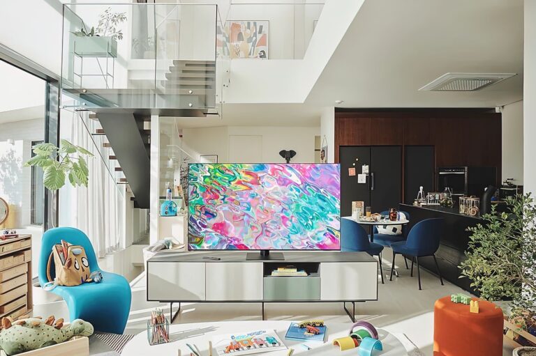 Nowoczesne wnętrze salonu z otwartą przestrzenią i jasnymi ścianami, w których widoczny jest telewizor Samsunga na niskiej szafce z kolorowym obrazem na ekranie, niebieskie fotel i sofa, zabawki na podłodze, drewniane schody z balustradą ze szkła, kuchnia w tle.