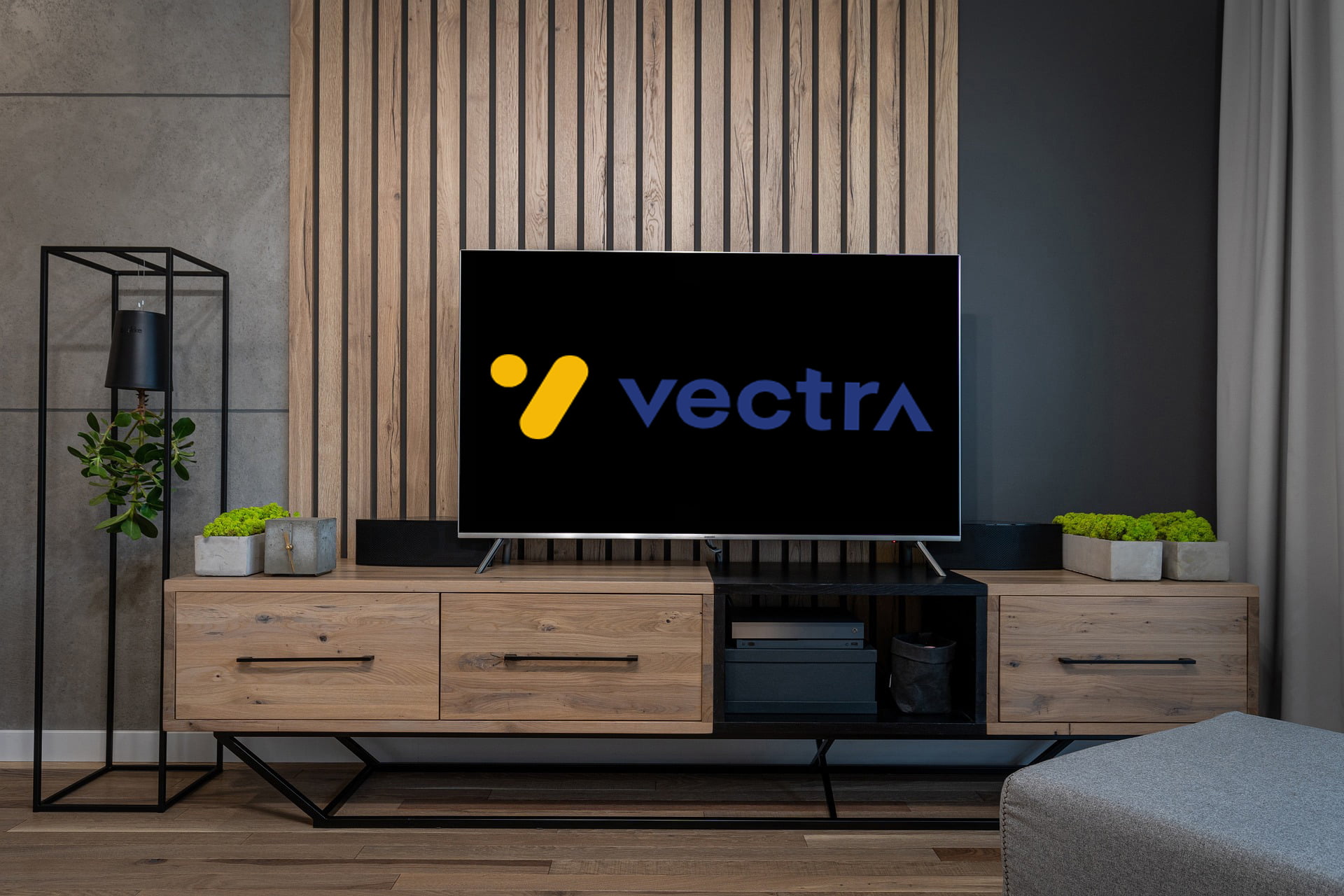 Wnętrze salonu z telewizorem na drewnianej komodzie wyświetlającym logo "vectra", obok doniczki z roślinami i lampa podłogowa, w tle ściana z drewnianymi panelami i szara zasłona.