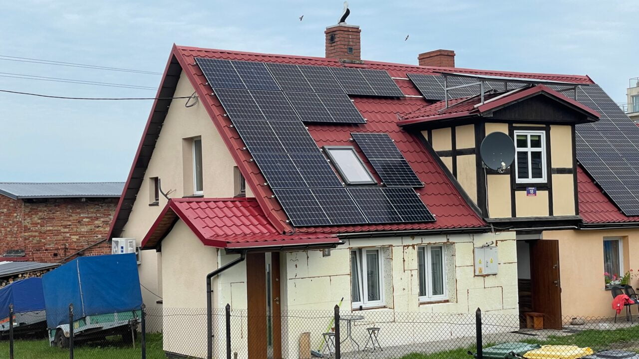 Dom jednorodzinny z czerwonym dachem i panelami słonecznymi, przed którym znajduje się ogrodzenie i przyczepa przykryta niebieską plandeką.