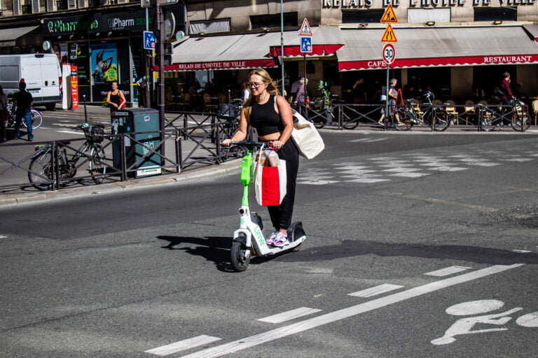 Kobieta jeździ na elektrycznej hulajnodze Lime na miejskim pasie rowerowym z torebką na ramieniu i zakupami.
