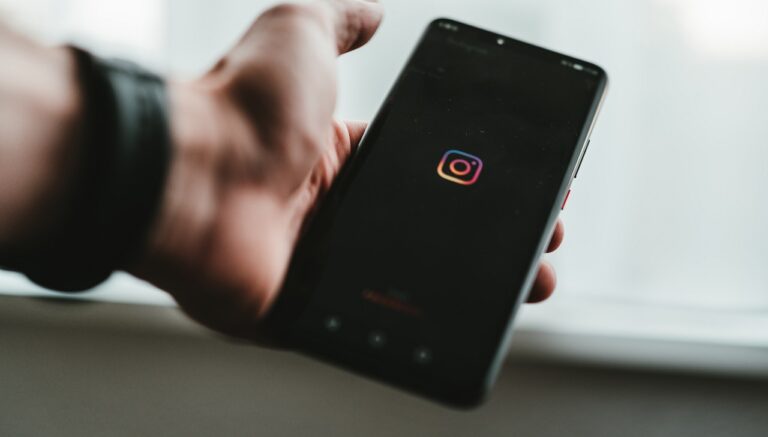 Zbliżenie na ekran smartfona trzymanego w prawej dłoni, wyświetlającego logo aplikacji Instagram na tle rozmytego.