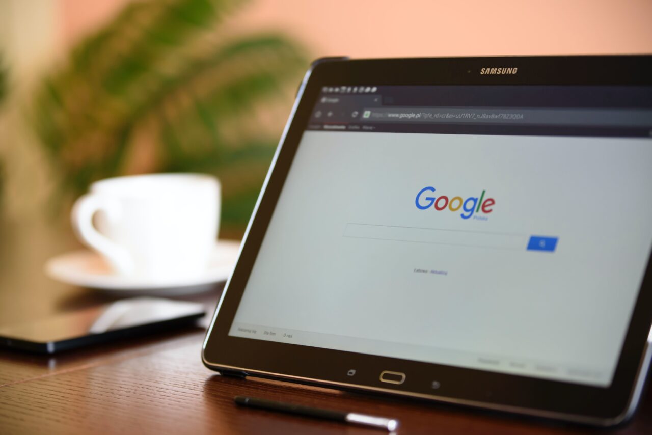 Tablet Samsung leżący na biurku z wyświetloną stroną główną wyszukiwarki Google, w tle rozmyta filiżanka kawy.