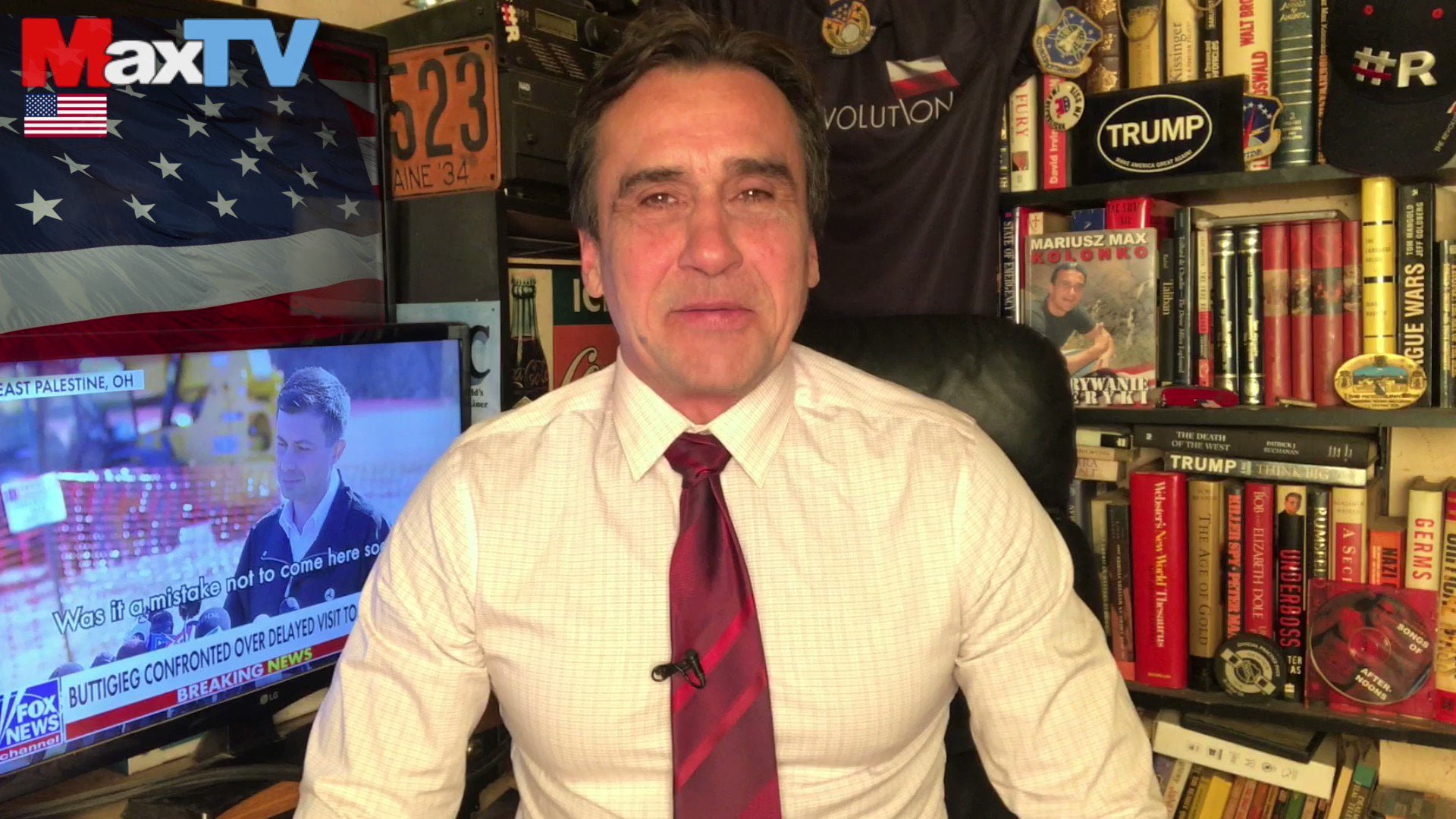 Mariusz Max Kolonko w białej koszuli i czerwonym krawacie stoi przed regałem z książkami i pamiątkami, a w tle widać telewizor wyświetlający kanał informacyjny oraz flagę USA.