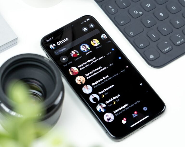 Smartfon z otwartą aplikacją do czatowania, wyświetlający listę rozmów, na biurku obok rozmazanego obiektywu aparatu i klawiatury.