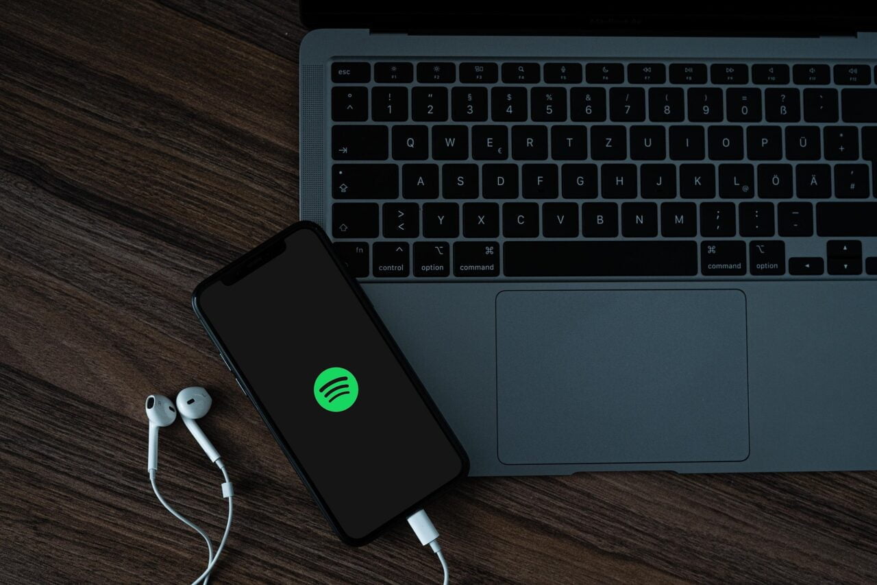 Smartfon z otwartą aplikacją Spotify połączony z białymi słuchawkami leży obok laptopa na drewnianym blacie.