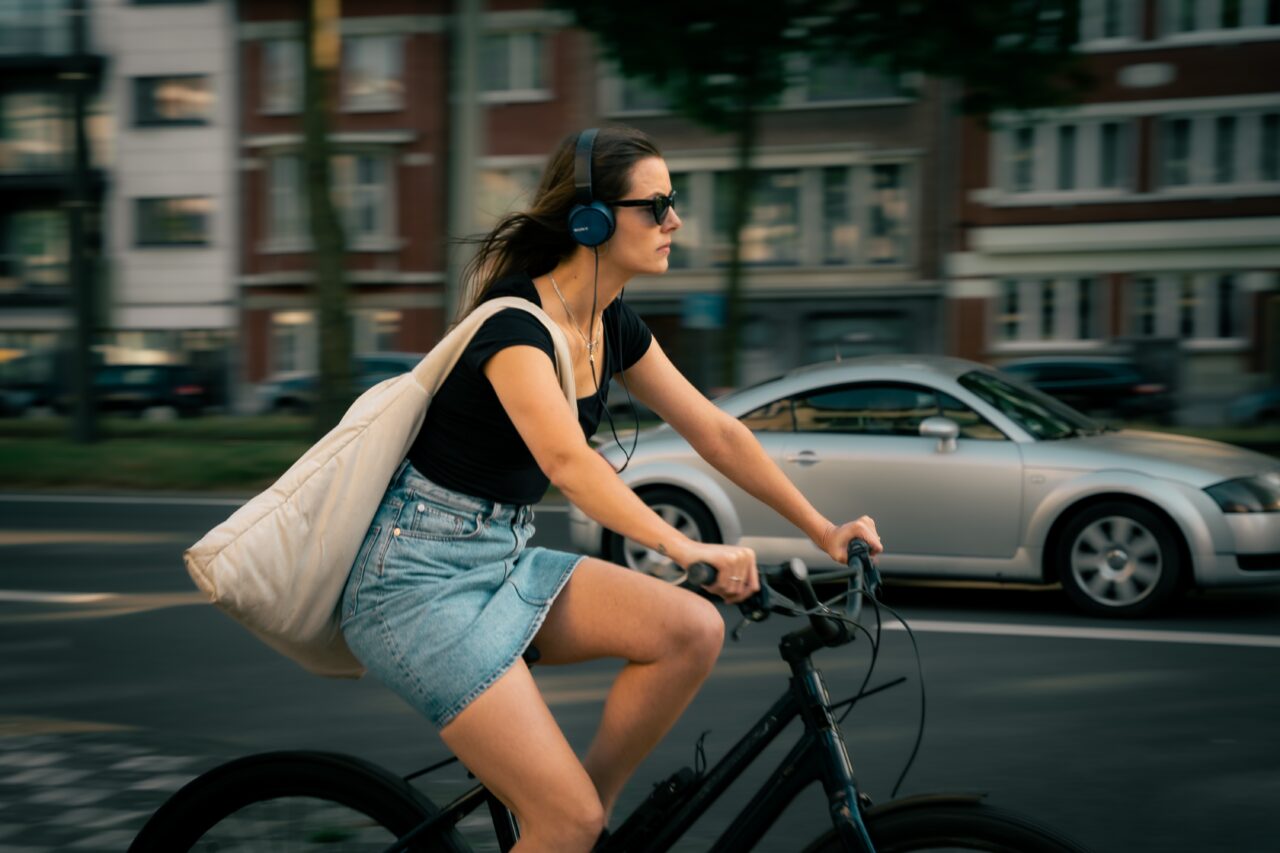 Kobieta w czarnej koszulce, dżinsowej spódnicy i słuchawkach na uszach jedzie na rowerze miejskim obok poruszających się samochodów.