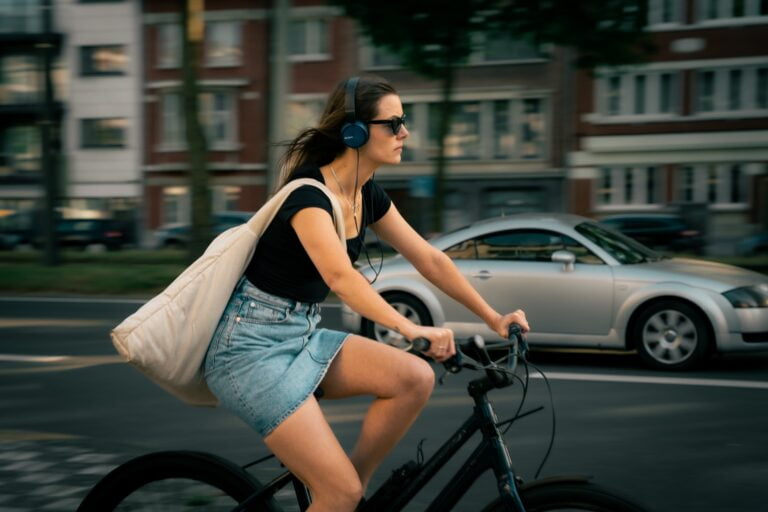 Młoda kobieta jedzie na rowerze po miejskiej ulicy, słuchając muzyki przez duże słuchawki nauszne, z tłem miejskich budynków i samochodu.