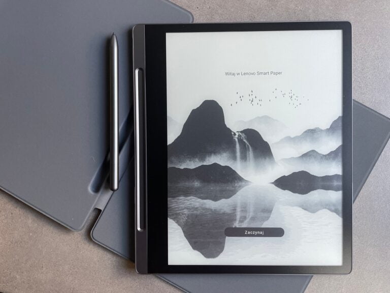 Elektroniczny czytnik dokumentów marki Lenovo z wyświetlaczem pokazującym górski krajobraz i napis "Witaj w Lenovo Smart Paper", położony na szarym laptopie z rysikiem obok.