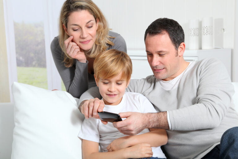 Rodzina z trójką osób: kobieta, mężczyzna i chłopiec patrzący na smartfona, siedzą razem na kanapie w jasnym pomieszczeniu.
