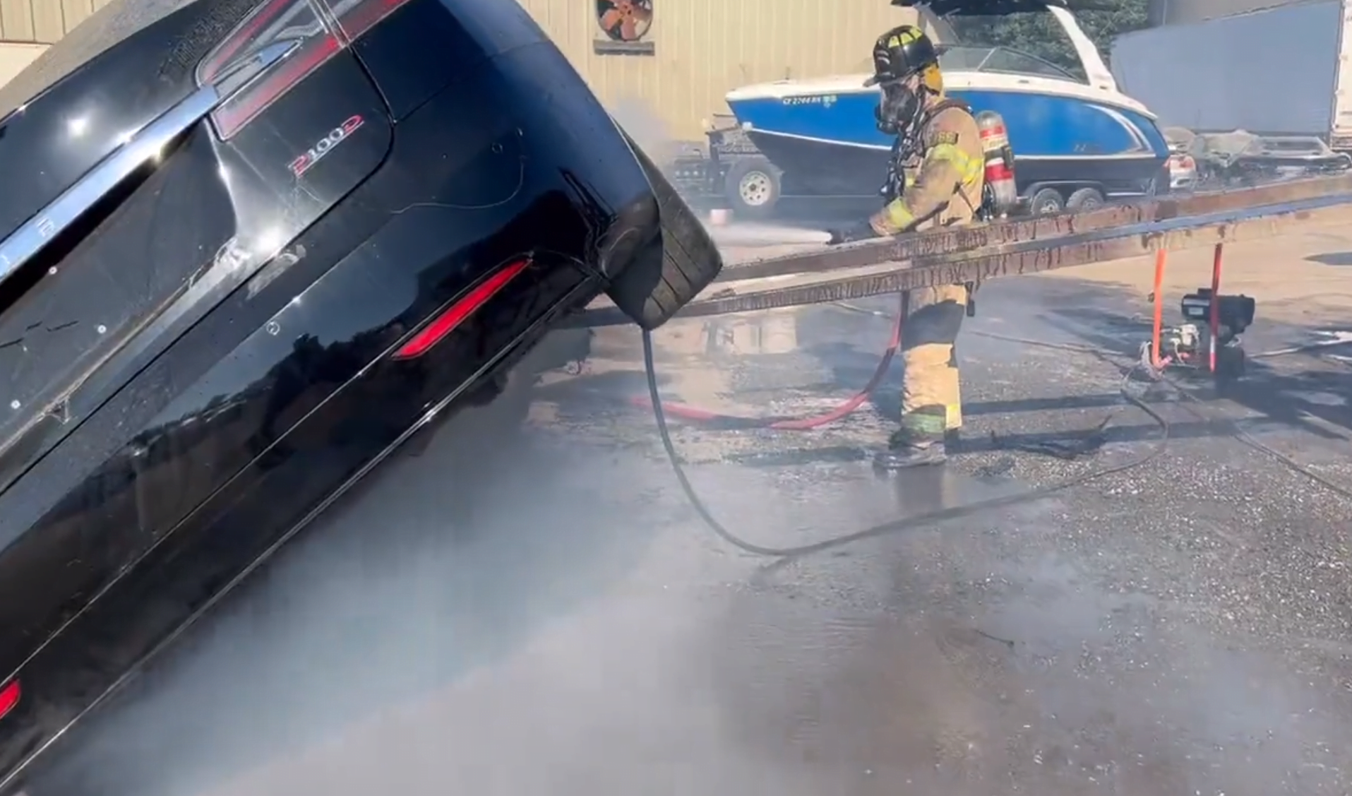 Strażak w pełnym ubioru gaśniczym trzyma wąż i gasi pożar uniesionego tylnego końca elektrycznego samochodu, z którego wydobywa się dym.