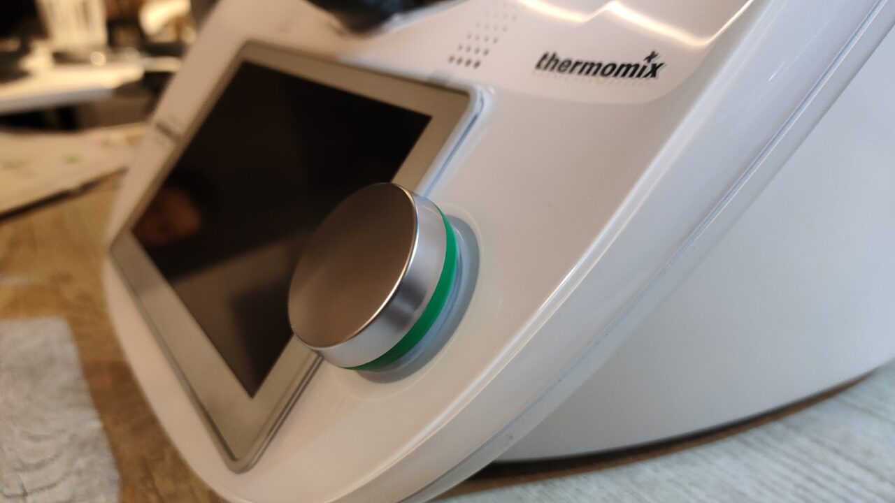 Robot kuchenny Thermomix TM6 z wyraźnym logo i pokrętłem sterującym z zielonym światłem na pierwszym planie, oraz częściowo widocznym ekranem dotykowym.
