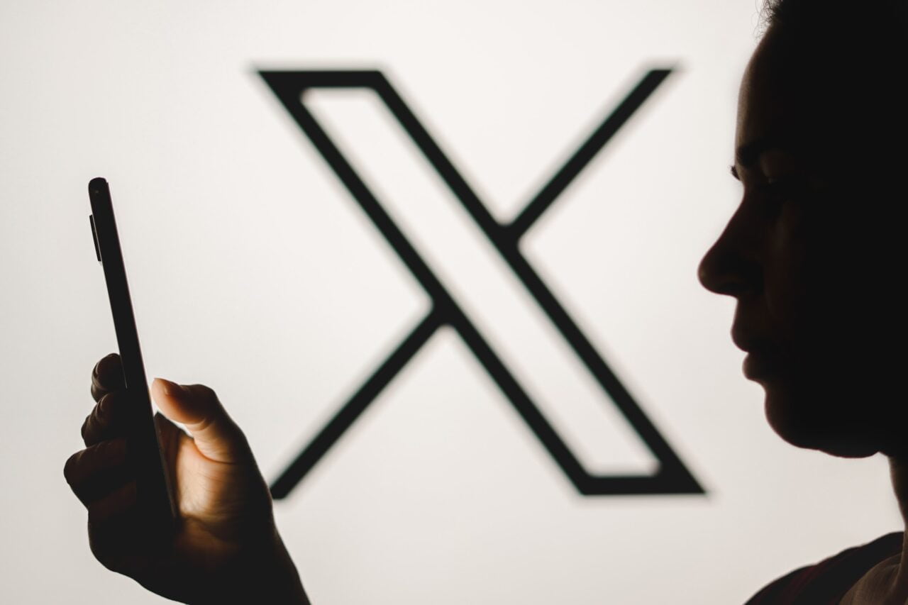 Mężczyzna patrzący w telefon, w tle czarne logo X na białym tle
