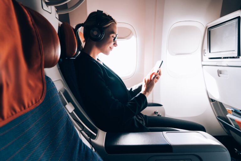 Kobieta w okularach i słuchawkach na uszach korzysta z telefonu komórkowego, siedząc przy oknie w samolocie.