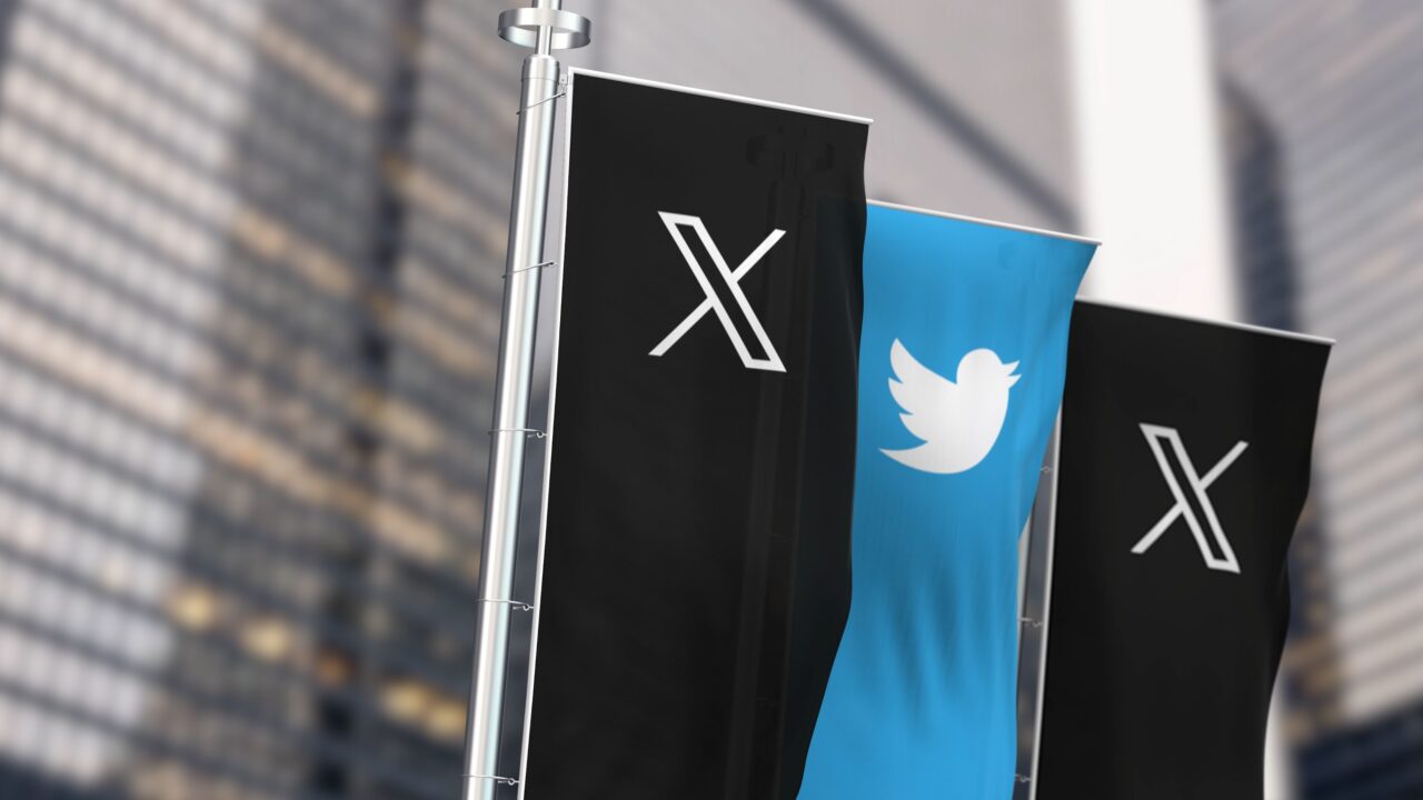 Trzy flagi (dwie X jedna Twitter) zawieszone na maszcie, środkowa z logotypem Twittera, z tłem w postaci wysokościowców.
