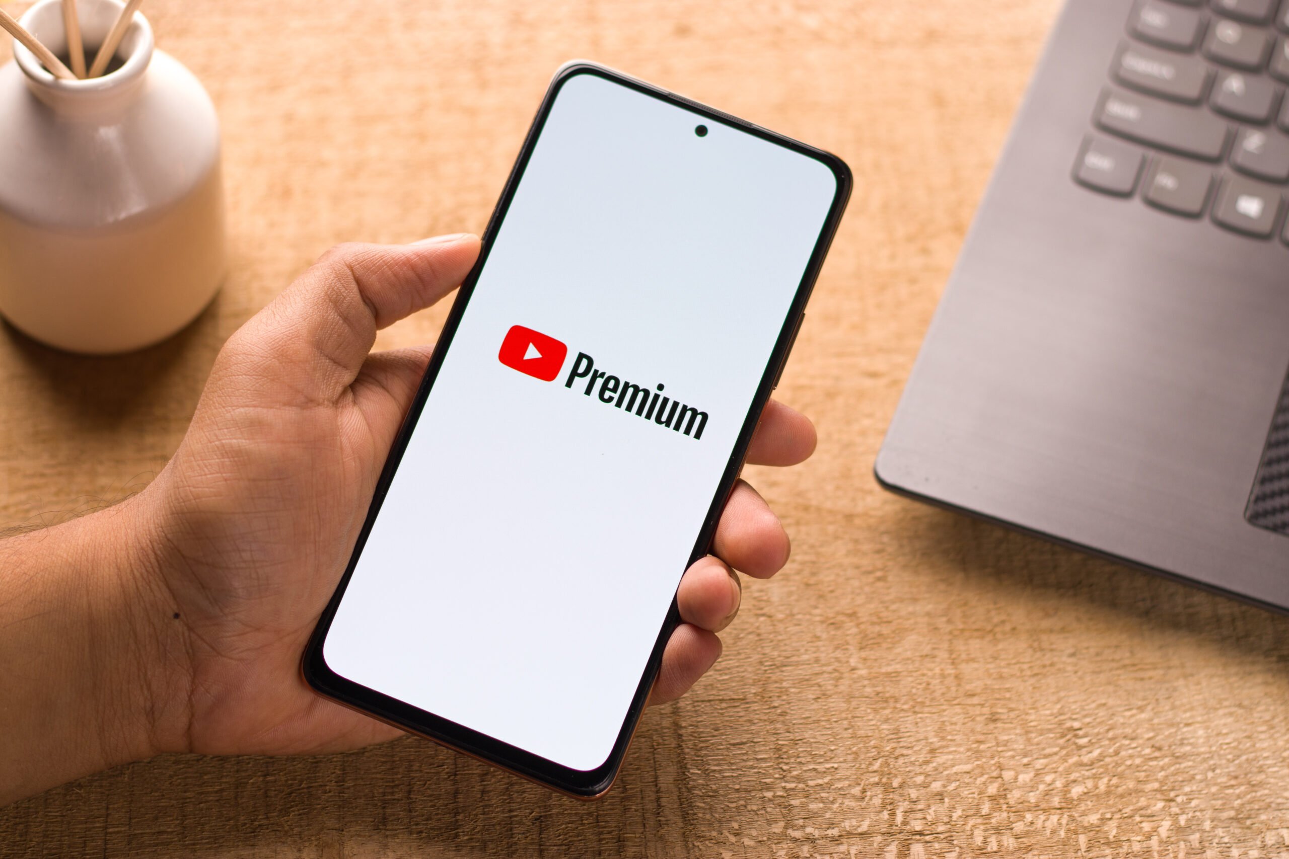 Smartfon z logo YouTube Premium na ekranie trzymany nad biurkiem i laptopem