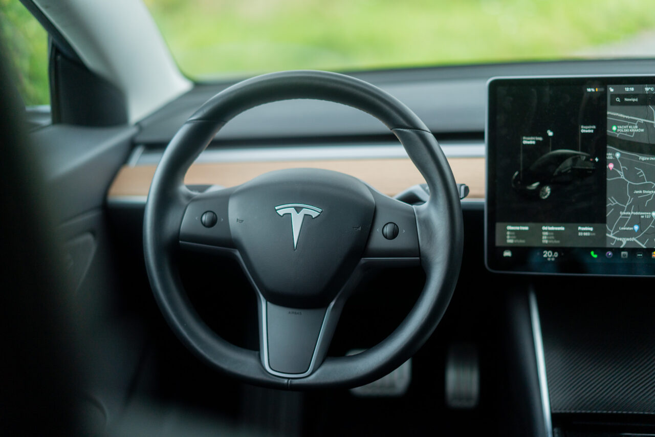 Kierownica samochodu elektrycznego marki Tesla z widocznym logo w centrum, w tle ekran systemu nawigacyjnego i kontrolnego.