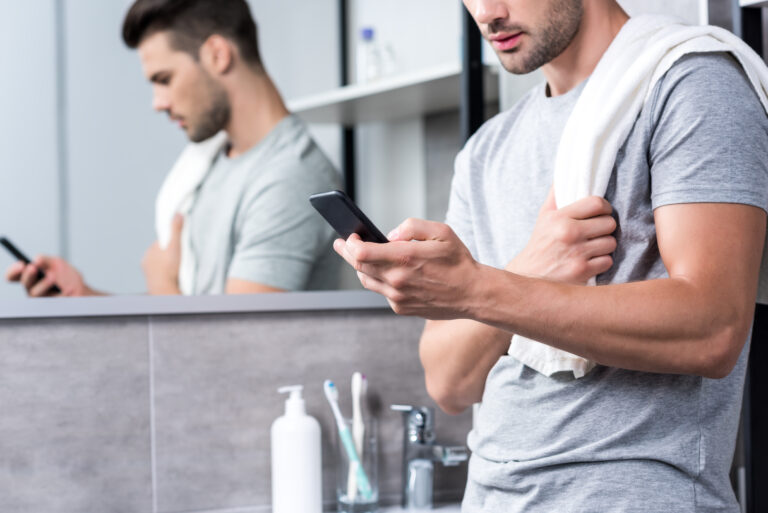 Młody mężczyzna w szarym t-shircie i z ręcznikiem na ramieniu używający smartfona przed łazienkowym lustrem.