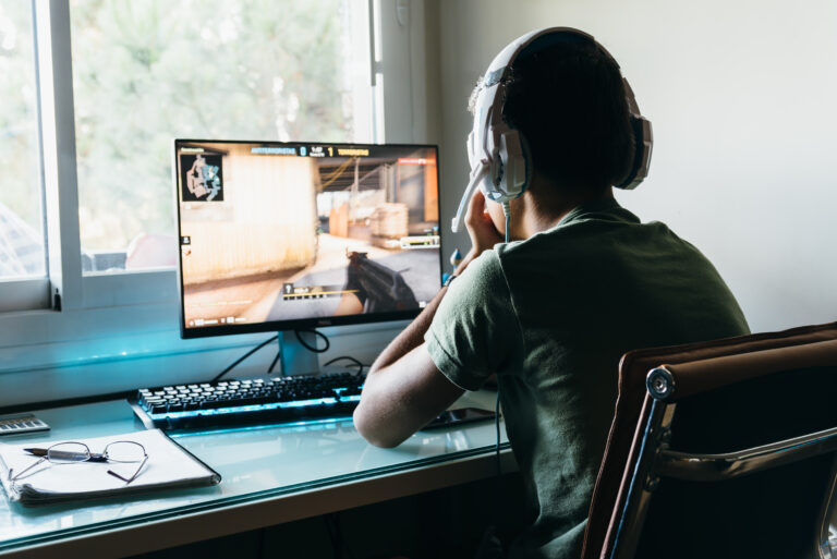 Osoba grająca w strzelankę na komputerze, z widokiem na monitor, nosząca słuchawki z mikrofonem, siedząca obok okna.