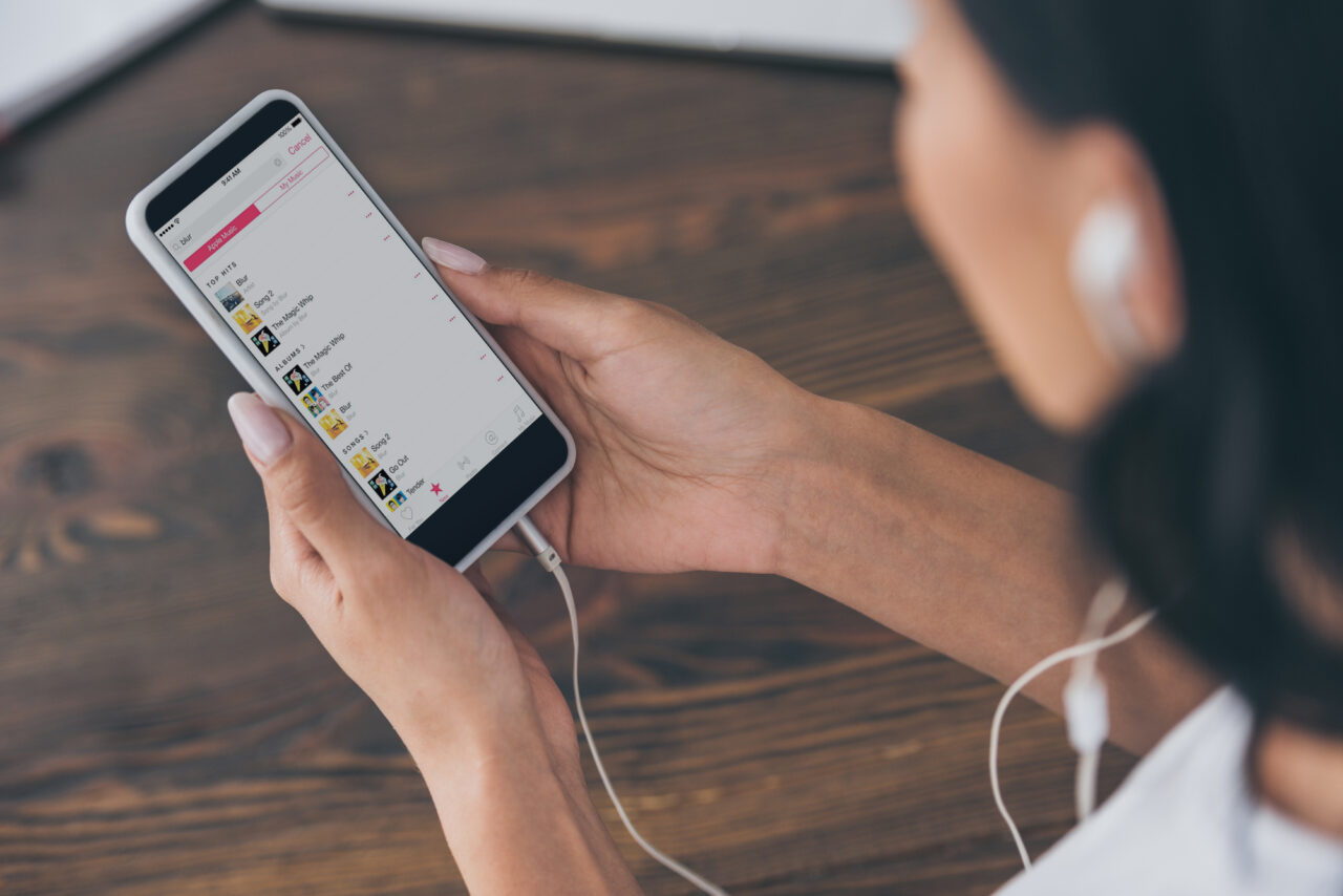 Apple Music vs Spotify. Kobieta trzyma smartfon z otwartą aplikacją do przesyłania muzyki, słuchając przez białe słuchawki douszne.