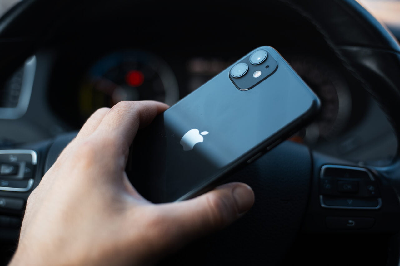Ręka trzymająca czarny iphone z logotypem Apple nad kierownicą samochodu.