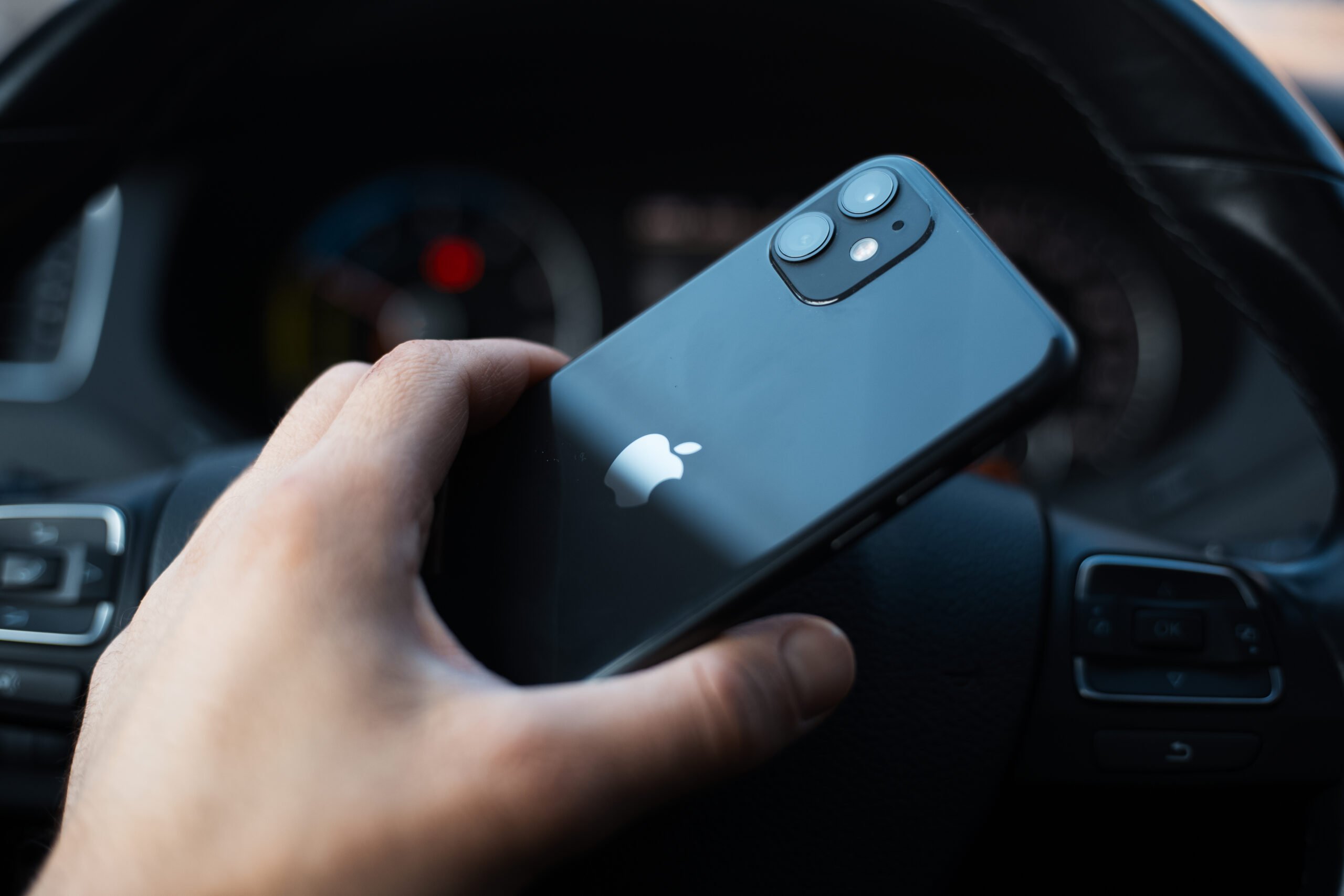 Ręka trzymająca czarny iphone z logotypem Apple nad kierownicą samochodu.