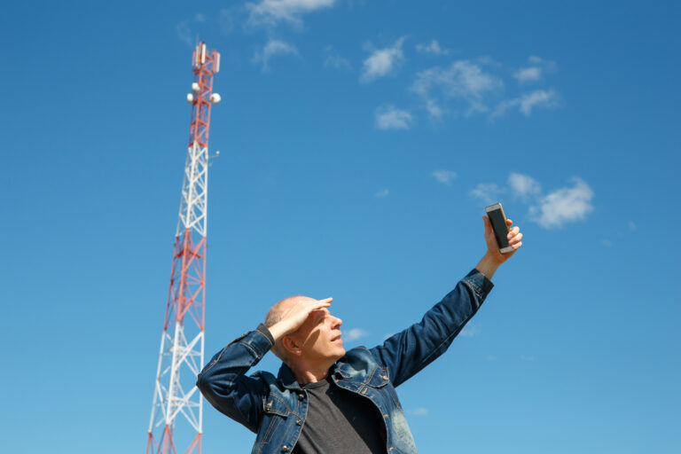 Mężczyzna w średnim wieku w jeansowej kurtce, trzymający smartfon i zasłaniający oczy ręką, patrzy w kierunku nieba z czerwonym radiowym masztem telekomunikacyjnym w tle.