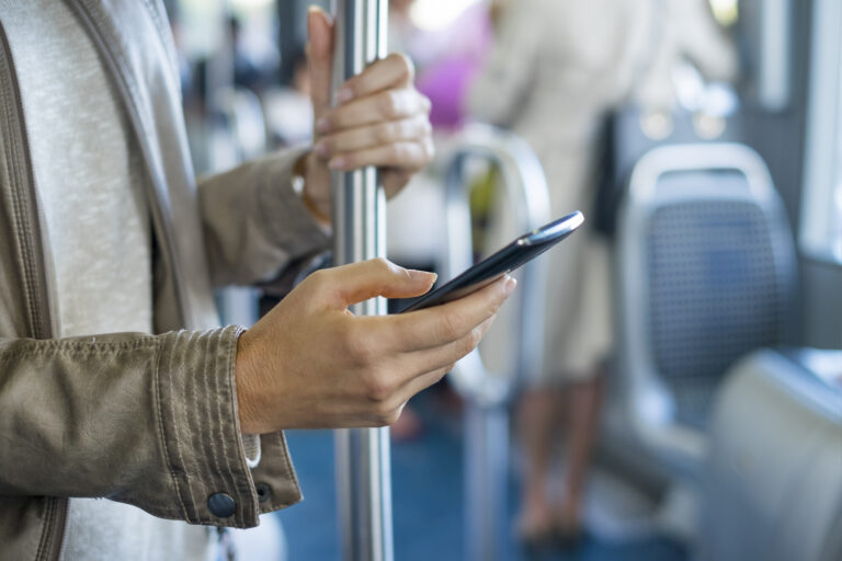 Osoba trzymająca smartfon w jednej ręce i poręcz w autobusie lub tramwaju w drugiej ręce, w tle widoczne siedzenia pojazdu komunikacji miejskiej.