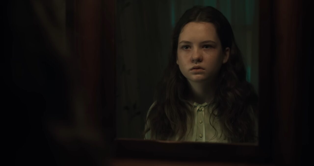 Kadr z filmu Egzorcysta: wyznawca. Młoda dziewczyna o poważnym wyrazie twarzy siedzi w półmroku, z troską patrząc przed siebie.