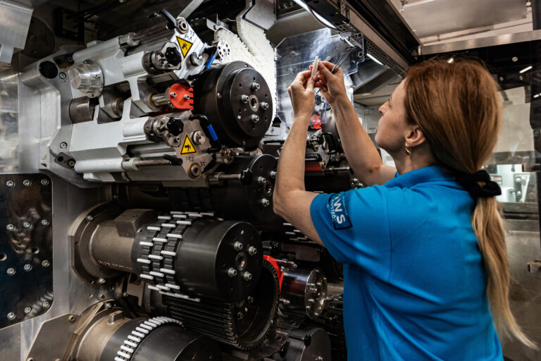 Kobieta w niebieskiej koszulce pracuje przy skomplikowanym przemysłowym urządzeniu z elementami mechanicznymi i elektronicznymi.