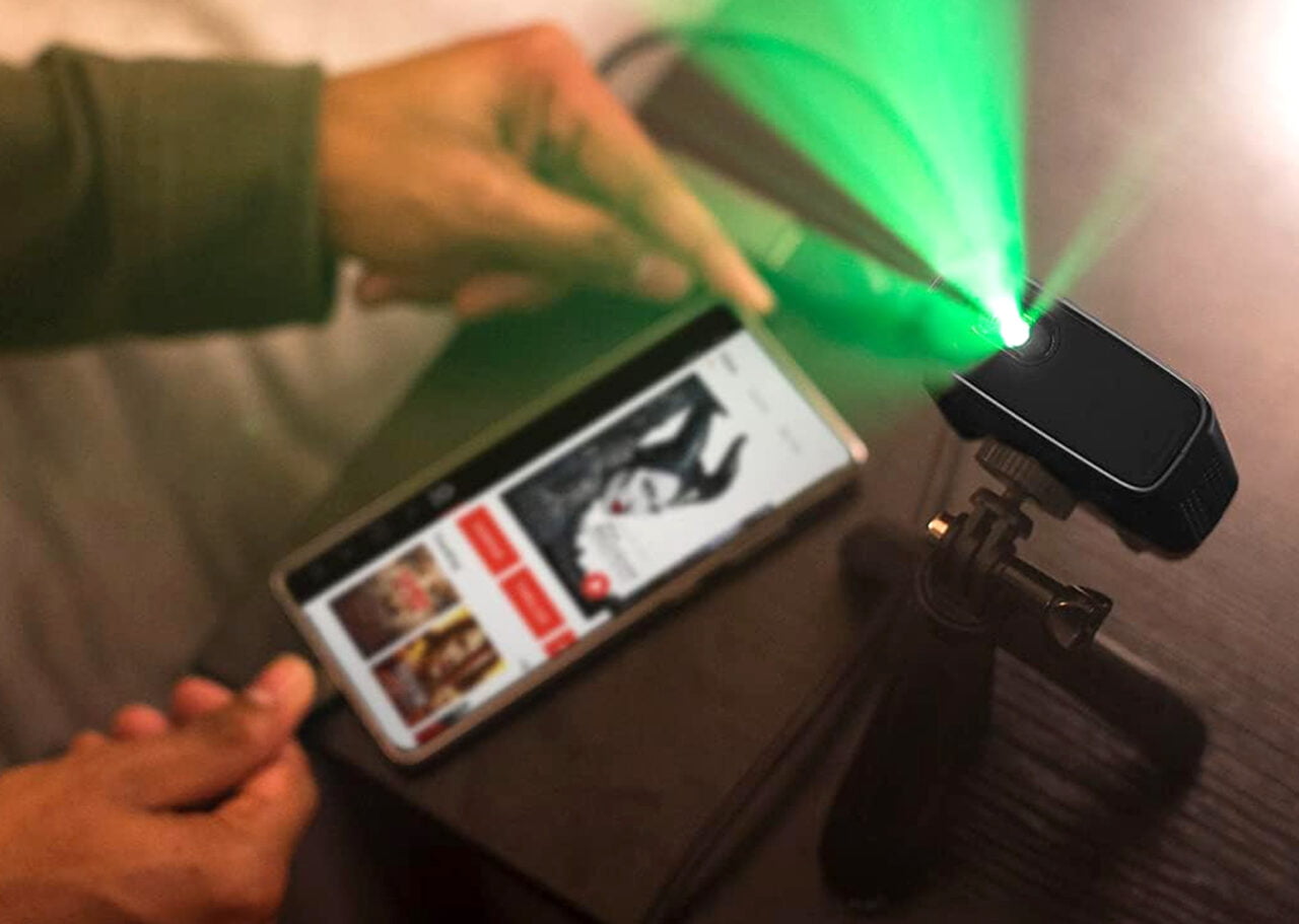 Projektor Philips PicoPix Micro Plus stojący na dokręcanej podstawie i wyświetlający zieloną poświatę. Obok mężczyzna trzymający smartfona 