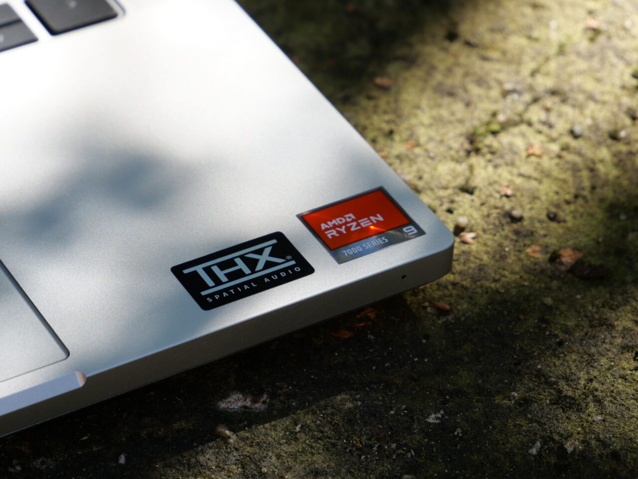 Część srebrnego laptopa z naklejkami THX Spatial Audio i AMD Ryzen 9 7000 Series na kamiennym podłożu.
