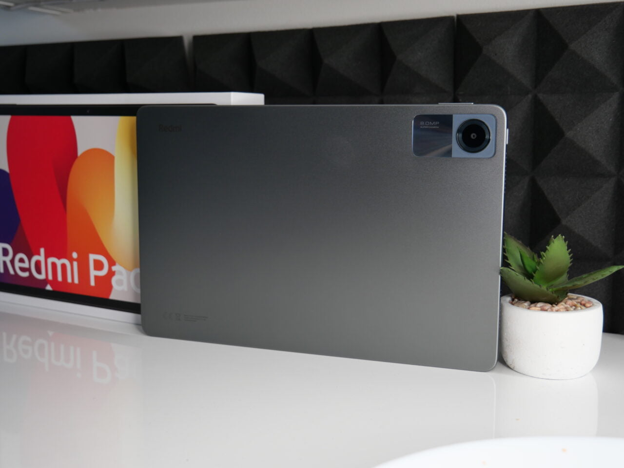 Tablet marki Xiaomi Redmi z tylnym aparatem fotograficznym, oparty o monitor z wyświetlonym logo Redmi, obok mała doniczka z sukulentem.