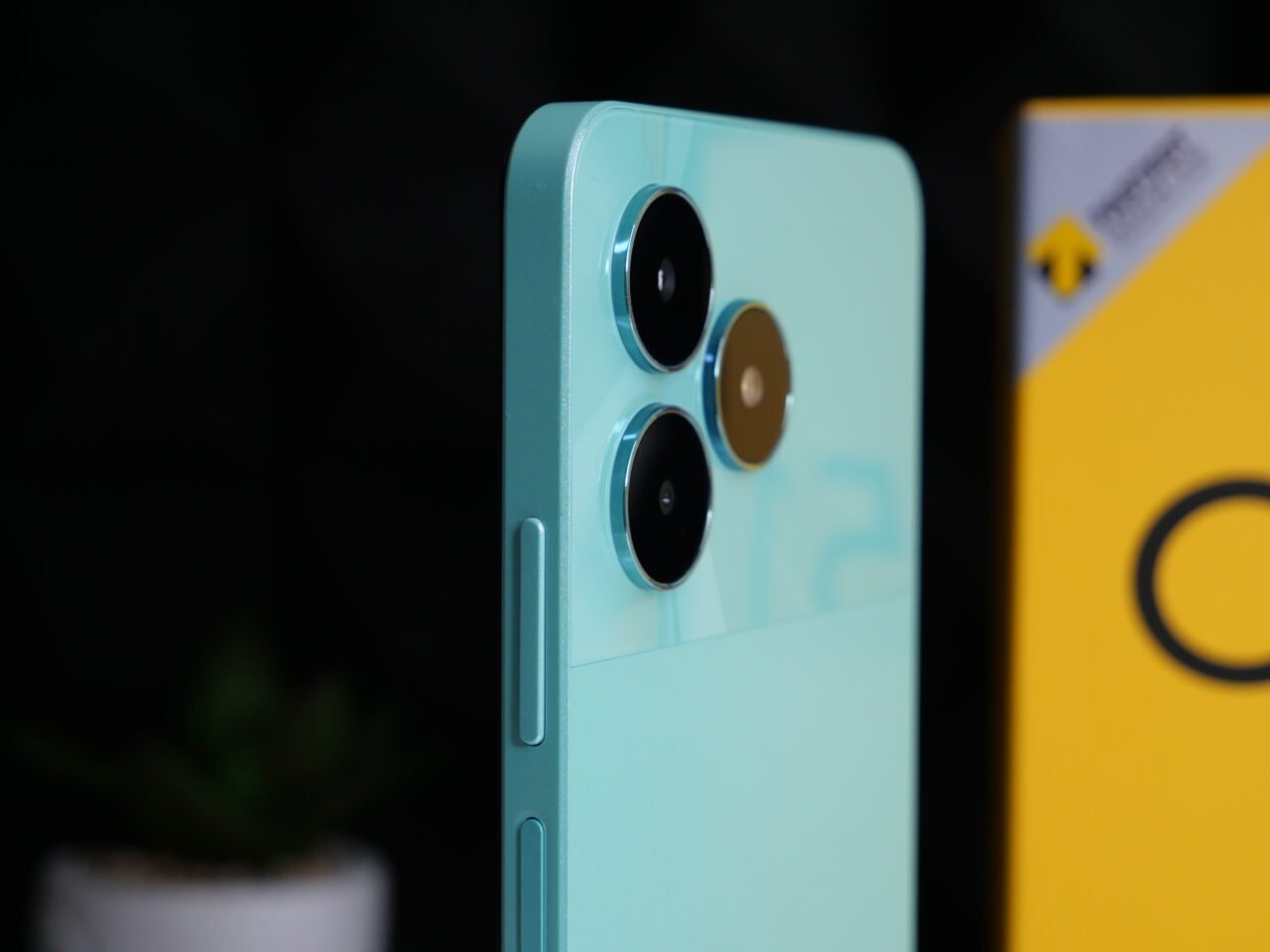 Smartfon w błękitnym kolorze z potrójnym aparatem i obiektywem w złotej obwódce, na tle rośliny i żółtego pudełka z logotypem.