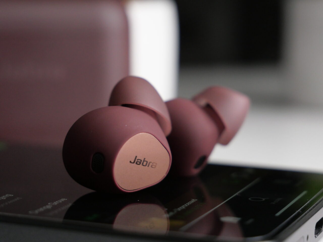 Zbliżenie bezprzewodowych słuchawek dousznych marki Jabra w kolorze bordowym, leżących na smartfonie.