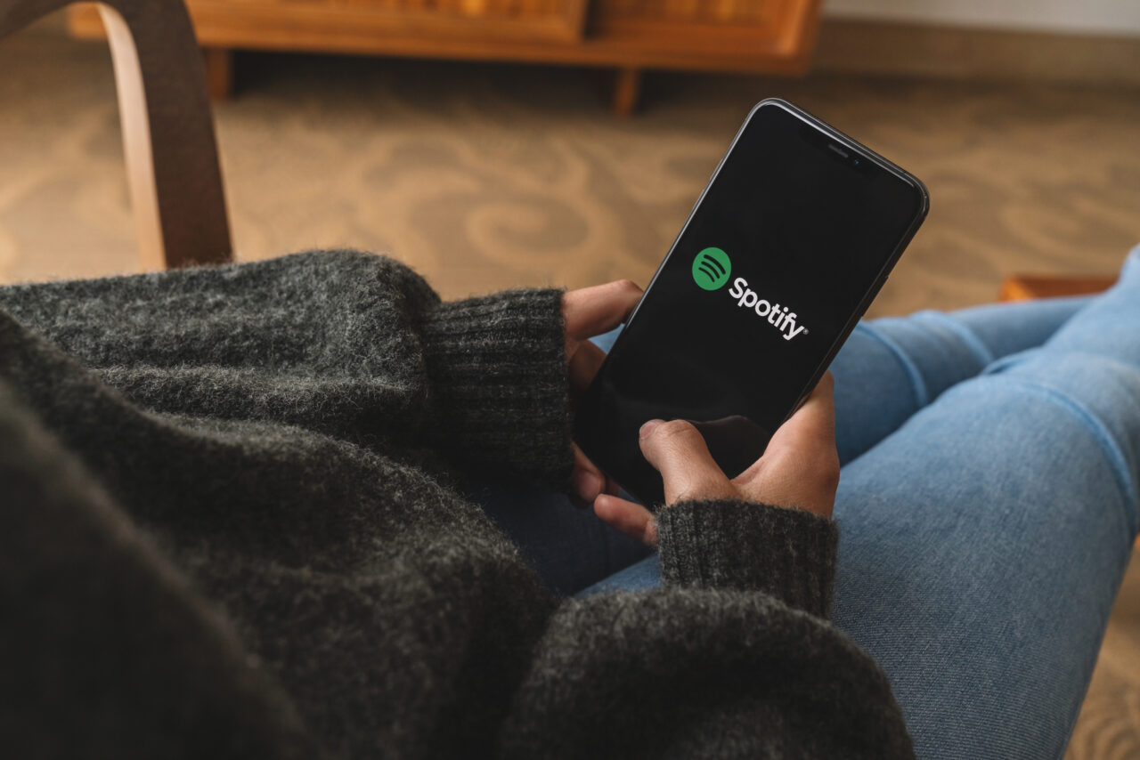 Uma pessoa segurando um smartphone com o logotipo do Spotify na tela, sentada em um sofá dentro de casa.