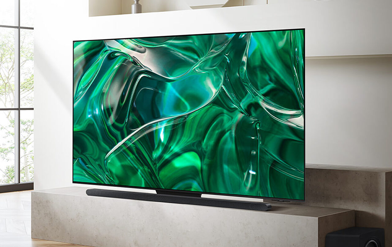 Duży telewizor Samsung z płaskim ekranem wyświetlający abstrakcyjne grafiki w odcieniach zieleni umieszczony na jasnym blacie w nowoczesnym salonie, pod telewizorem znajduje się soundbar, a po prawej stronie widoczny jest niewielki subwoofer.