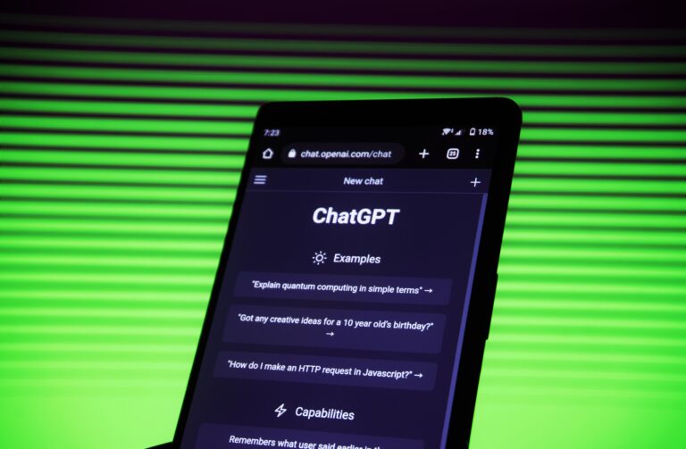 Smartfon wyświetlający ekran czatu ChatGPT na stronie chat.openai.com z przykładowymi pytaniami, na zielonym, pionowo prążkowanym tle.