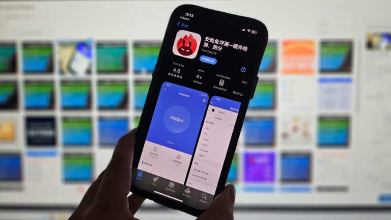 Smartfon trzymany w dłoni przed rozmytym tłem z ekranami komputerowymi, wyświetla chińskojęzyczną aplikację mobilną.