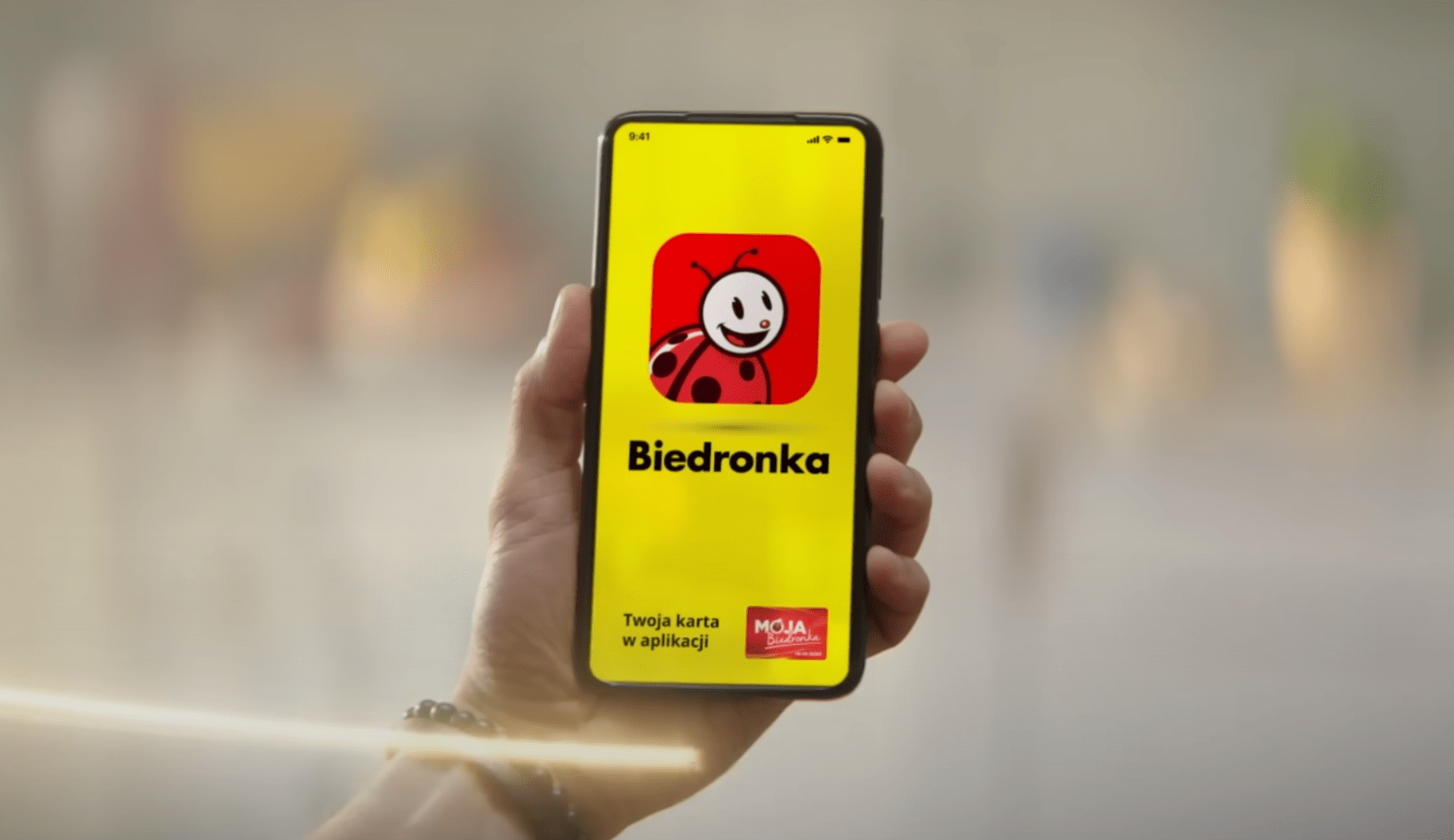 Ręka trzymająca smartfon z ekranem wyświetlającym aplikację Biedronka z logo w postaci czerwonego biedronki na żółtym tle oraz napisem "Twoja karta w aplikacji MOJA Biedronka".
