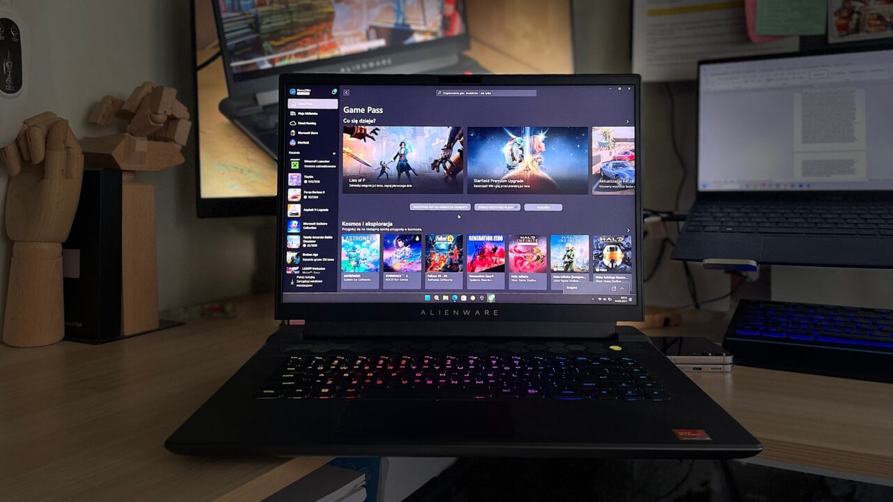 Laptop gamingowy Alienware na biurku z otwartym ekranem pokazującym interfejs biblioteki gier Game Pass, z klawiaturą podświetloną na różne kolory w tle.