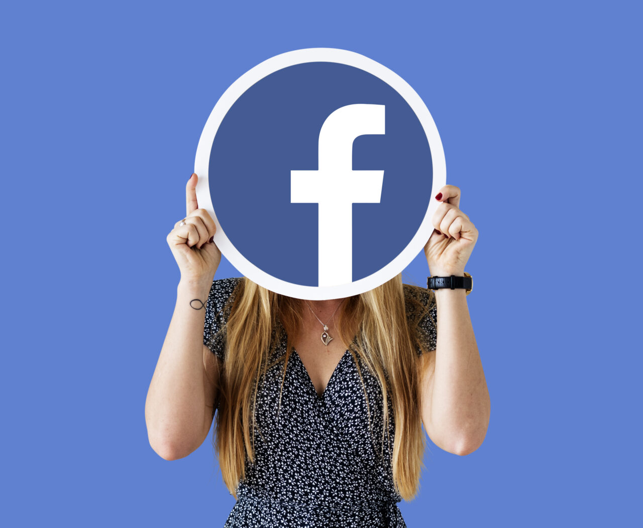 Ilustracja do tekstu Facebook nie działa. Osoba trzymająca duże logo Facebooka przed twarzą na niebieskim tle.
