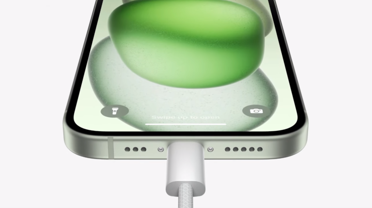 Smartfon z zieloną tapetą i podłączonym białym przewodem ładowania USB-C, pokazując komunikat "Swipe up to open" na ekranie blokady.