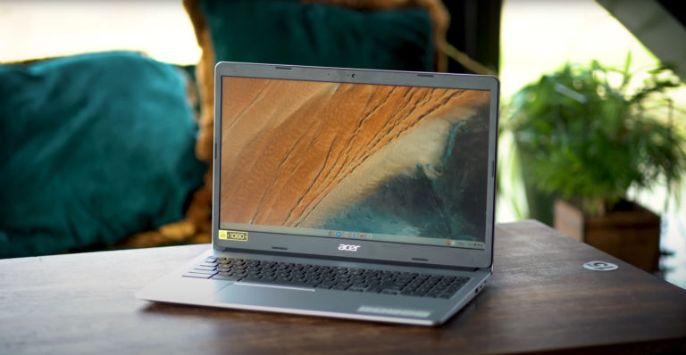 Laptop firmy Acer otwarty na drewnianym stole, z widocznym górzystym krajobrazem na ekranie, w tle roślina i poduszka na krześle.
