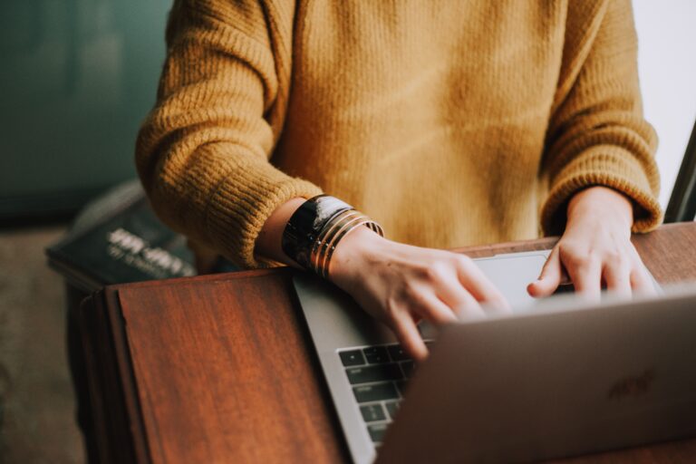 Osoba w żółtym swetrze pracująca na laptopie, oparta o drewniane biurko, z widocznymi rękami i biżuterią na nadgarstku.