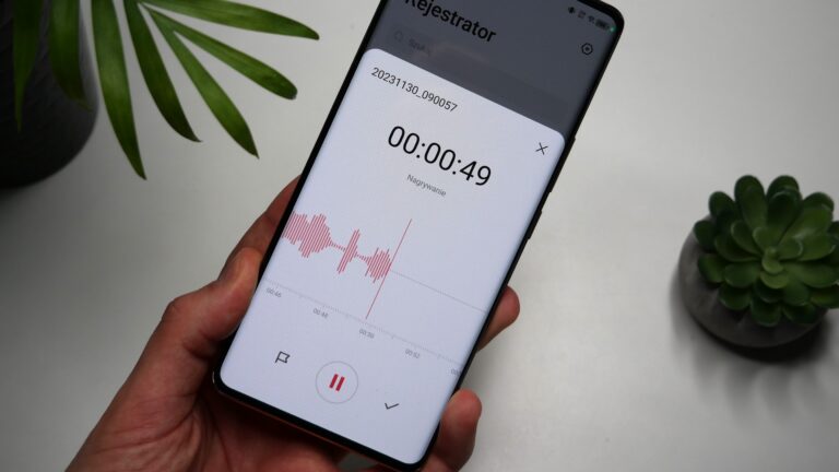 Smartfon trzymany w dłoni z otwartą aplikacją dyktafonu, pokazujący falę dźwiękową nagrania i czas trwania 00:00:49. Na biurku rośliny doniczkowe.